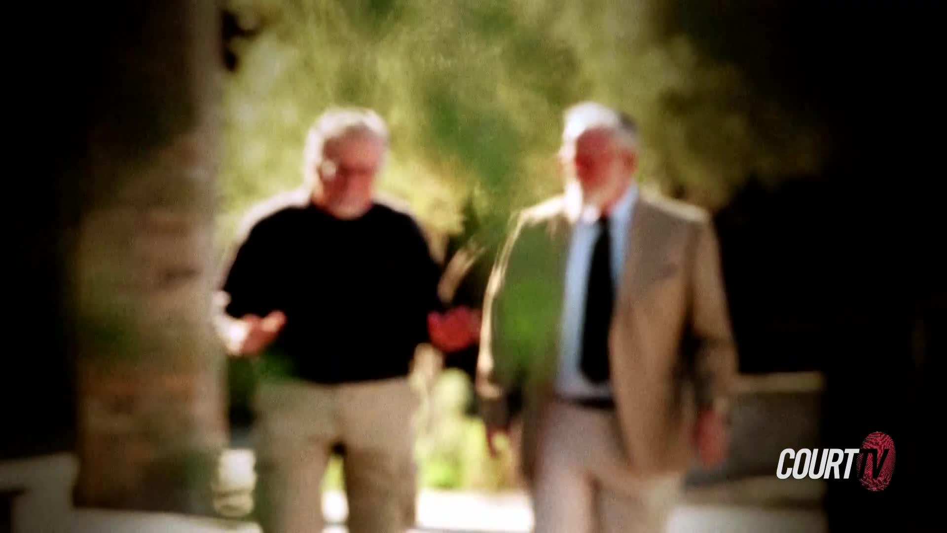 Two men walk toward me, one in a black shirt and khakis, the other in a suit jacket and tie. They appear to be discussing something important, perhaps related to a case being covered by Court TV.