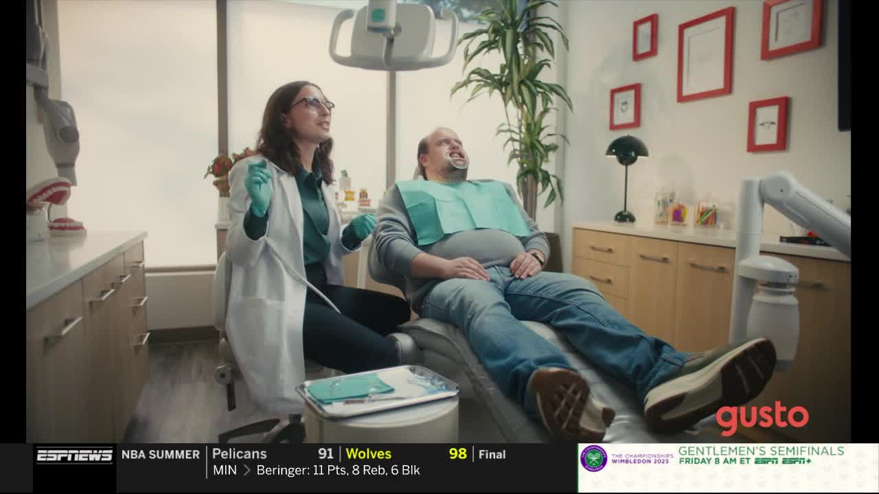 A dentist, wearing a white coat and gloves, leans over a patient in a dental chair. The patient has his mouth open, a bib around his neck, and is staring up at the dentist.
