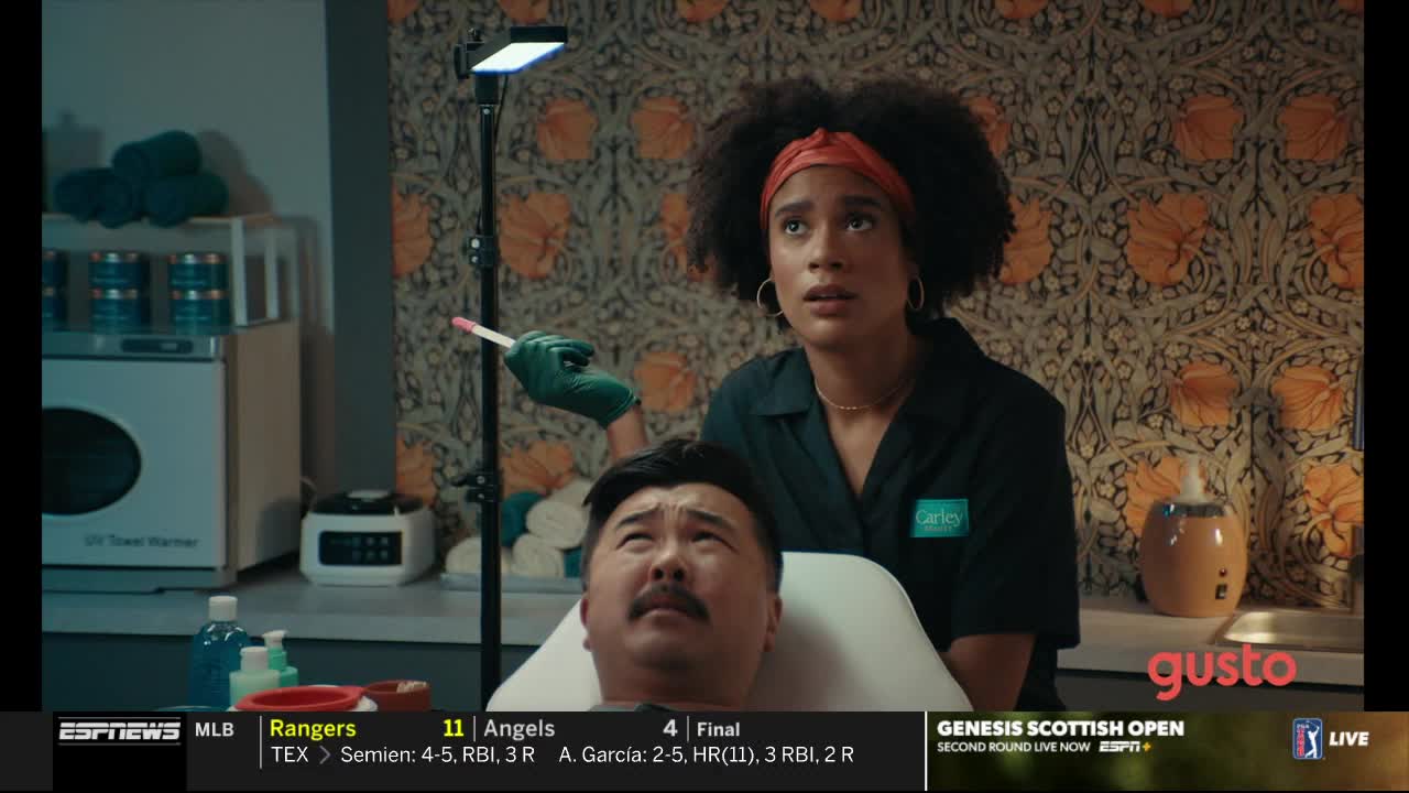 A woman in a black shirt holds a pink stick near a man's face, her expression a mix of concern and anticipation. The man is reclined, looking up as if awaiting a procedure.
