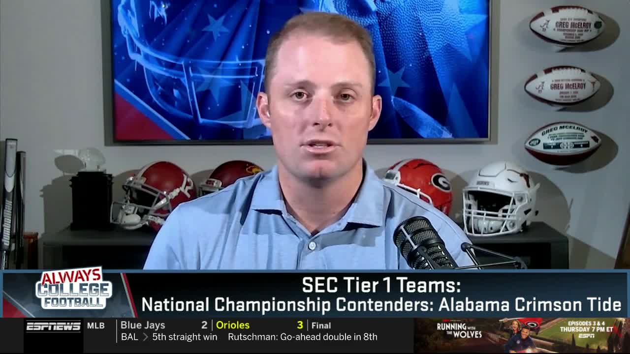 A man in a light blue shirt speaks into a microphone, discussing college football on ESPNews. Behind him, football helmets and signed footballs are displayed, along with a graphic listing Alabama as a championship contender.
