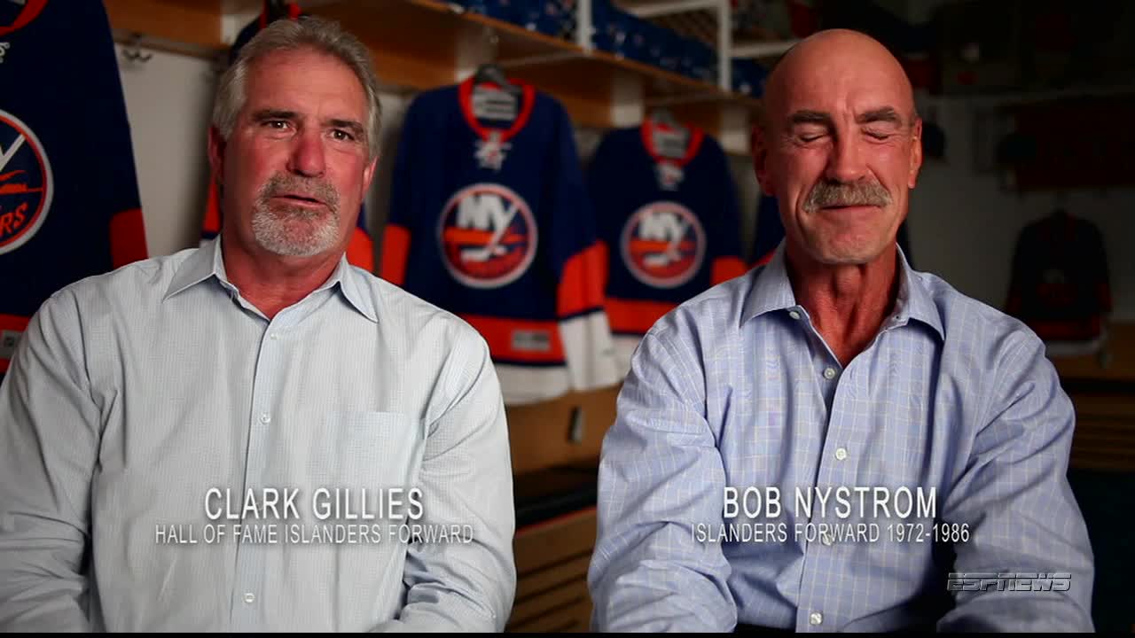 Two men, Clark Gillies and Bob Nystrom, sit side-by-side, both wearing button-down shirts. Behind them, the blue and orange jerseys of the New York Islanders hang on display.

