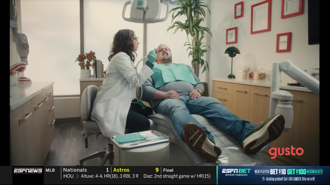 A dentist, wearing a white coat, works on a patient's teeth, who is reclined in a chair. The scene is a commercial on ESPNews, with baseball scores displayed at the bottom.
