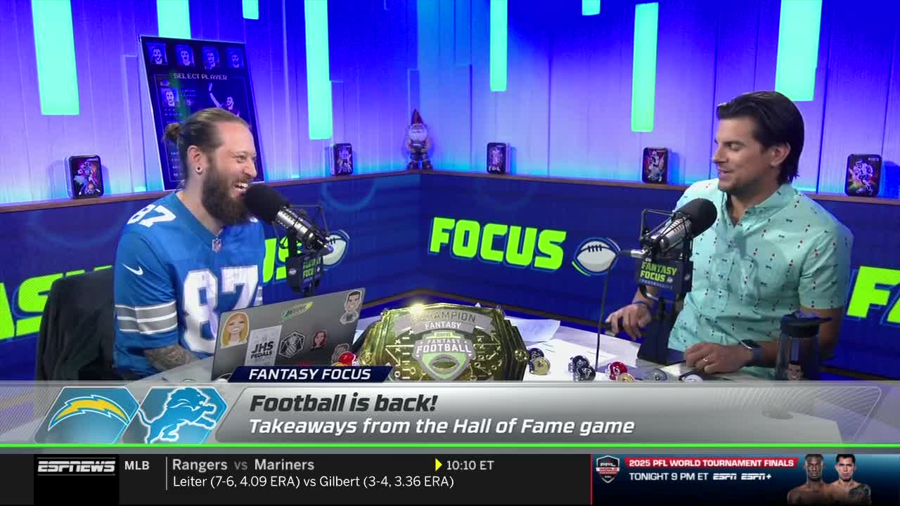 Two men are seated at a desk, microphones in front of them, on an ESPNews set. The man on the left, wearing a Detroit Lions jersey, is laughing, while the other man smiles.
