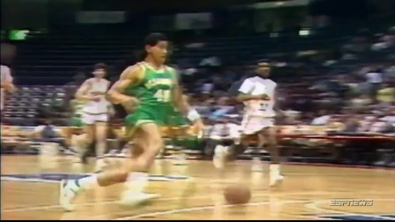 A player in a green jersey dribbles the ball toward the basket, kicking up a small cloud of dust. Another player in white is running to catch up, while others are further behind.
