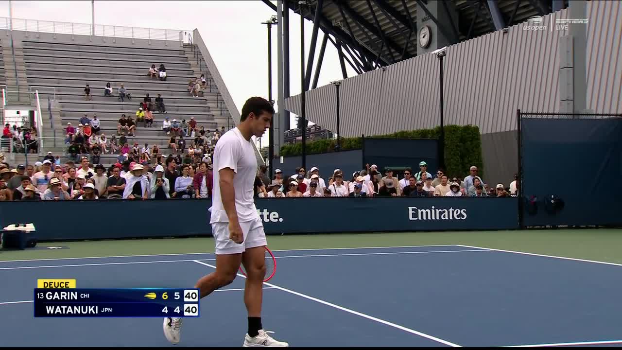 The tennis player in white walks toward the back of the court, racket in hand, after a point. The scoreboard on the screen displays the score of the match, which is being broadcast on ESPNews.
