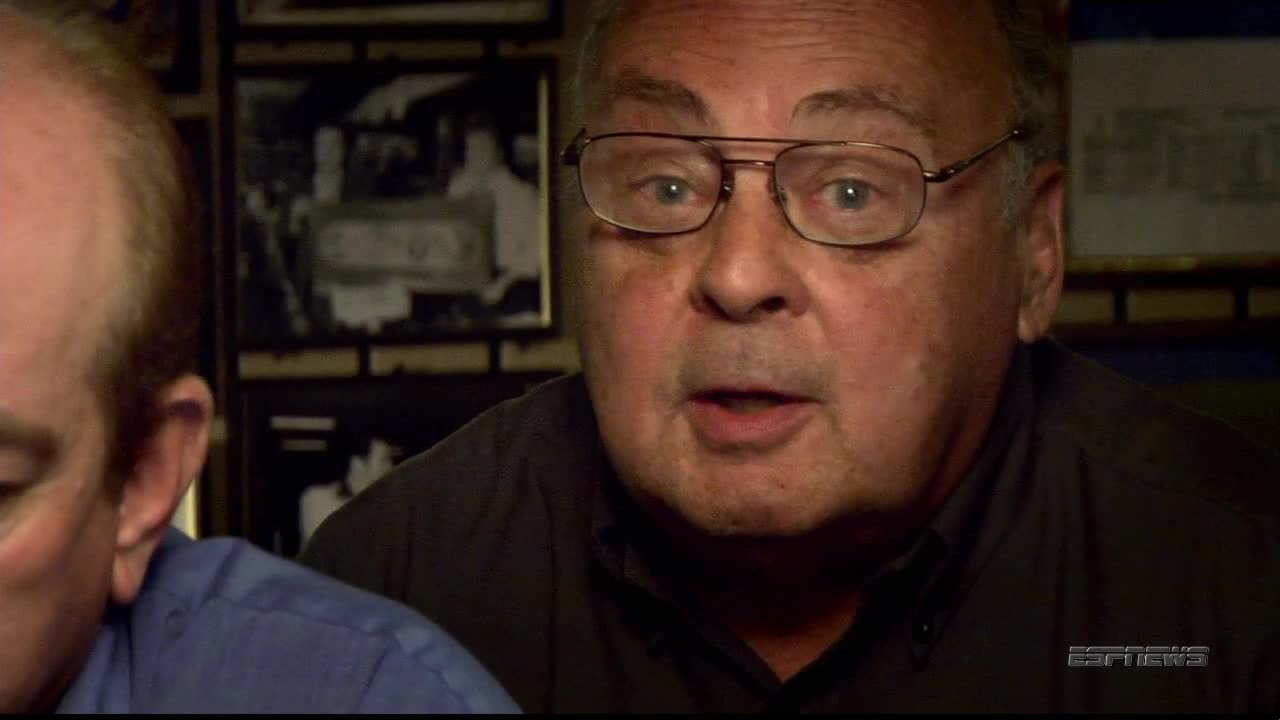 The man in glasses is speaking, his mouth open, and his eyes wide.  Beside him, another man's head is visible, both men are in a dimly lit room.
