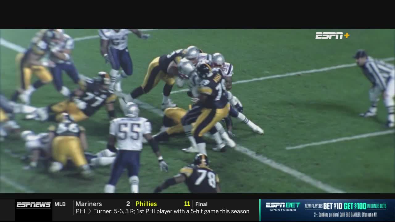 Steelers and Patriots players are tangled in a pile near the sideline, the yellow and black jerseys clashing with the blue and silver. A referee in black and white stripes watches the scrum unfold on the ESPNews broadcast.
