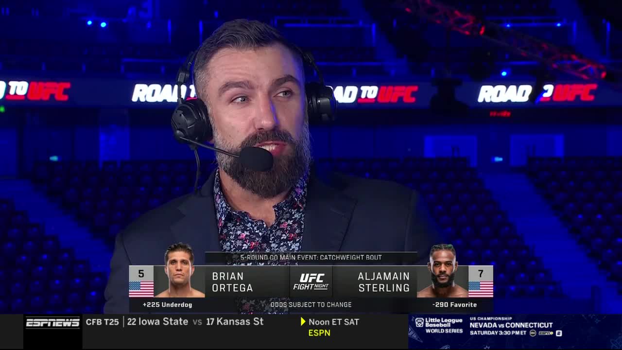 A man with a beard and a headset speaks into a microphone, likely on ESPNews. Graphics below him show the names of two fighters, Brian Ortega and Aljamain Sterling, for a UFC fight.
