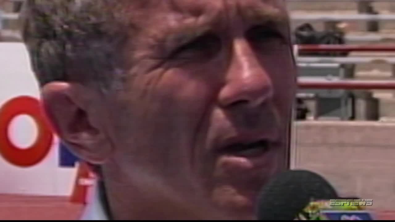 The man is speaking into a microphone, his face lit by the sun. ESPNews is visible on the screen, and a large red "O" is behind him.
