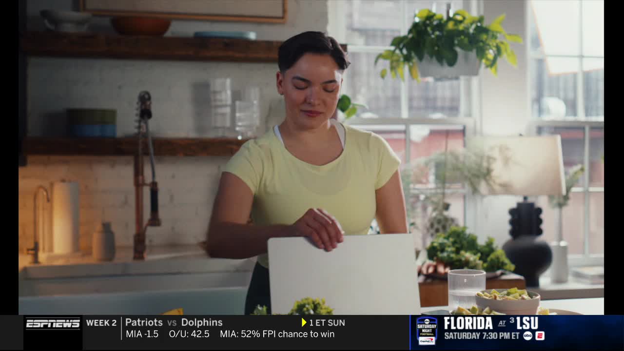 A woman in a yellow shirt looks down at a laptop in a sunlit kitchen. The ESPNews banner at the bottom of the screen shows a football game is on.
