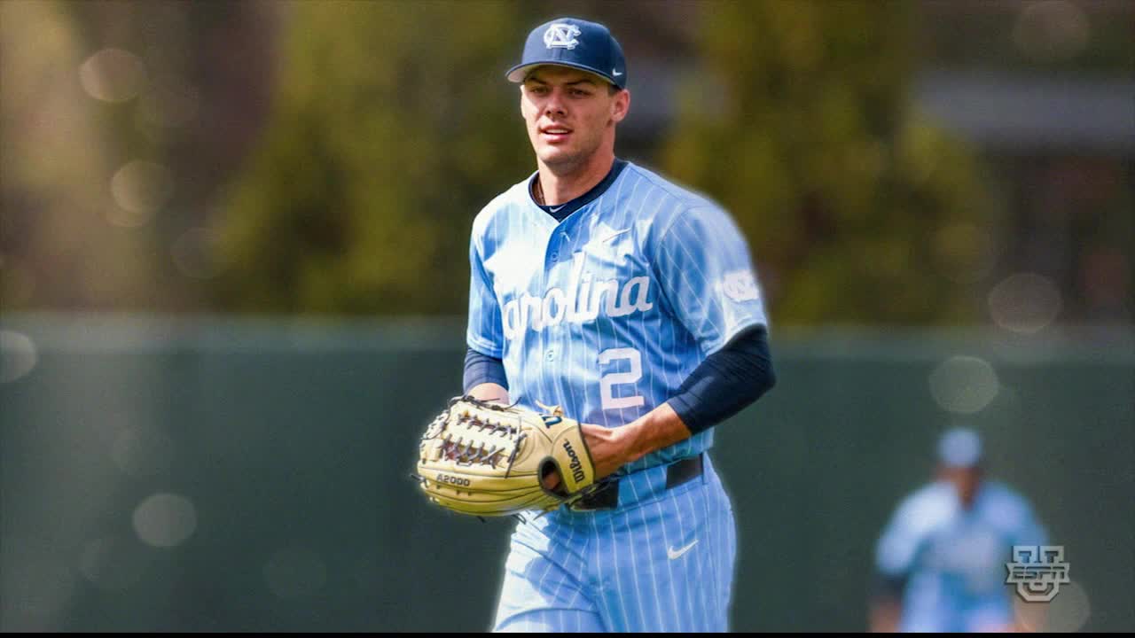The baseball player, wearing a Carolina jersey, stands on the field, glove in hand. He looks towards the camera, a slight smile on his face.
