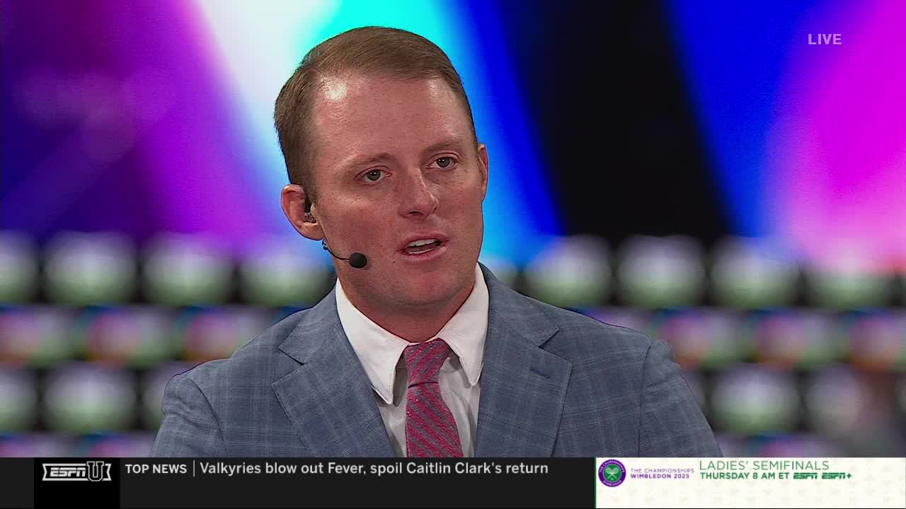 A man in a suit jacket and a pink tie is speaking, a microphone clipped to his ear. Behind him, a backdrop of blurred colors provides a backdrop for the ESPNU broadcast.
