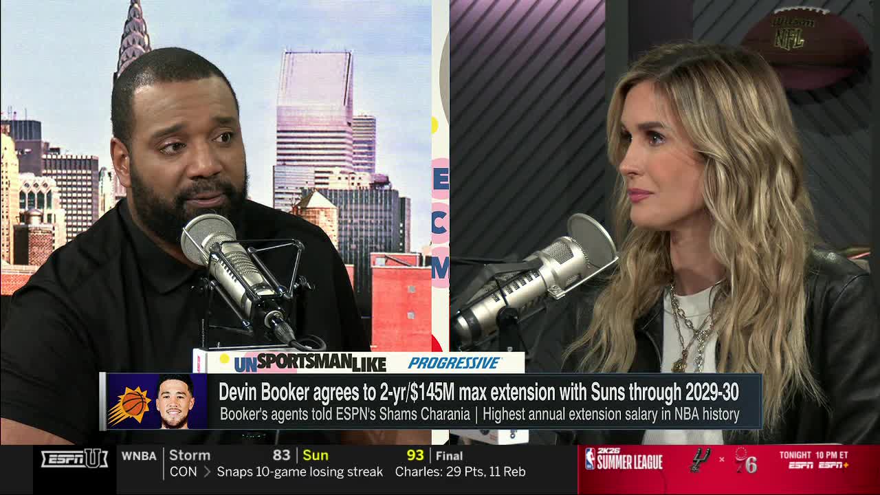 Two people are seated at a table, facing each other with microphones in front of them. The man on the left is speaking, while the woman on the right listens intently.

