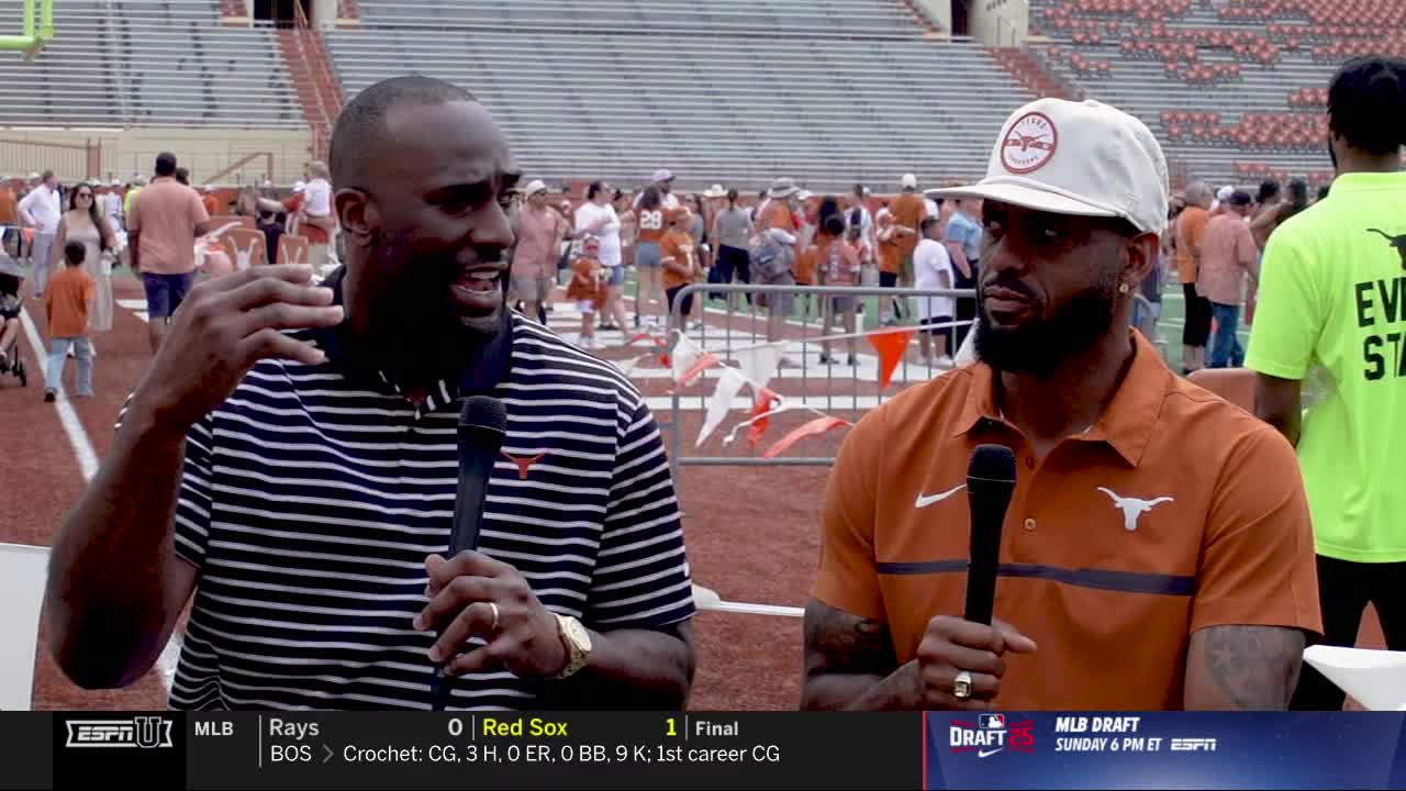 Two men, each holding a microphone, are speaking on the sidelines of a football field. Behind them, a crowd of people in orange and white are milling about.
