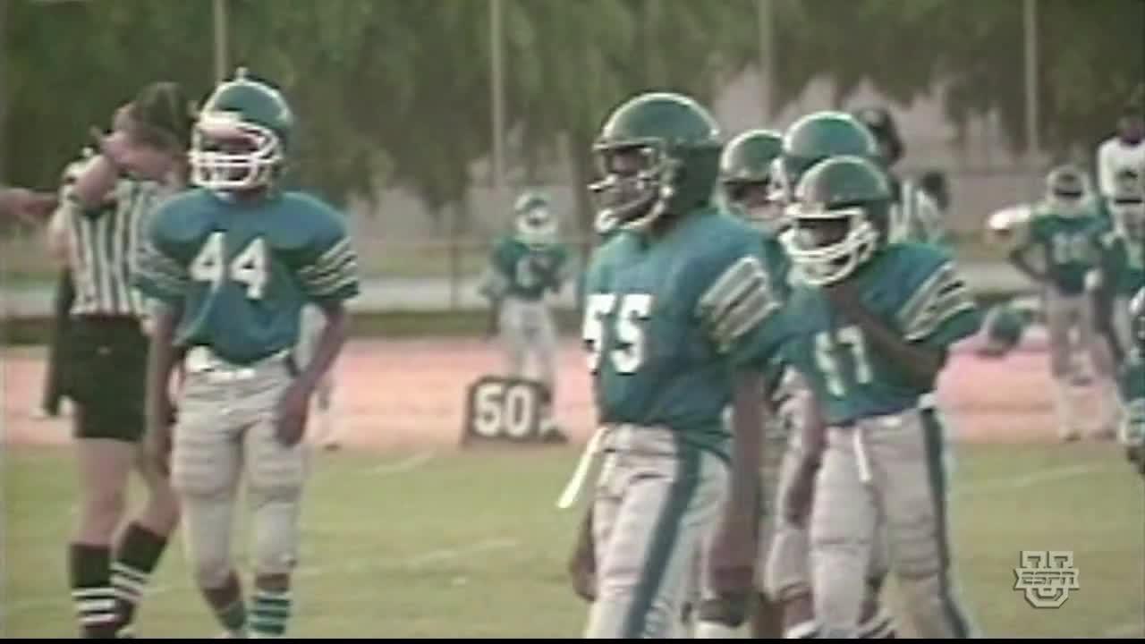 Players in teal jerseys and white pants stand on the field, helmets on, waiting. The referee in black and white stripes looks on as the 50-yard line marker looms in the background.
