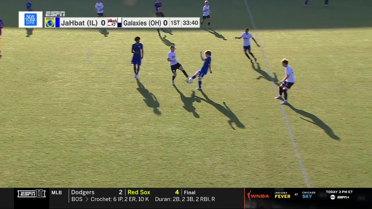 A player in blue kicks the ball toward the right, while an opposing player in white tries to intercept. Other players spread out across the green field, casting long shadows in the sunlight.
