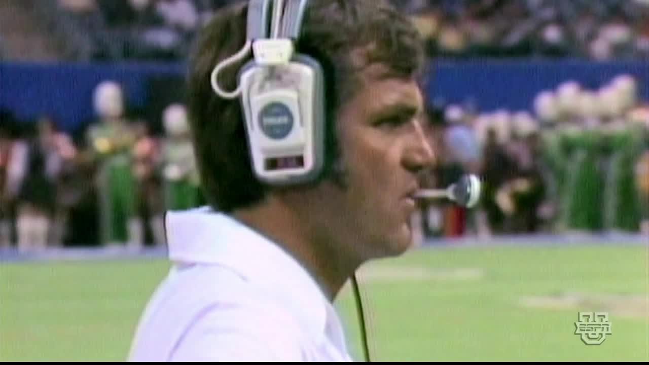 The coach, wearing a headset, is focused on the field, the microphone near his mouth. The green of the field is visible, and the ESPNU logo is in the corner.
