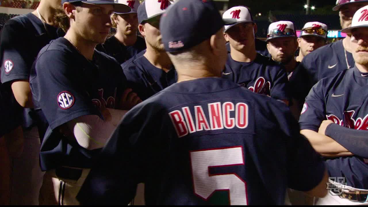 Coach Bianco, in his jersey, is facing his team, speaking. The players, in navy uniforms, listen intently, some with arms crossed.
