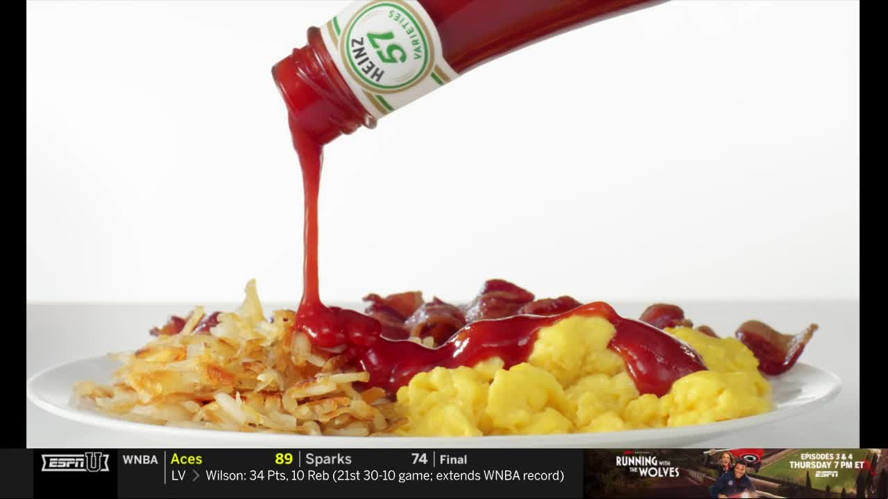 A bottle of Heinz ketchup pours a thick, red stream onto a plate of scrambled eggs, hash browns, and bacon. The food sits on a white plate, and the background is plain.
