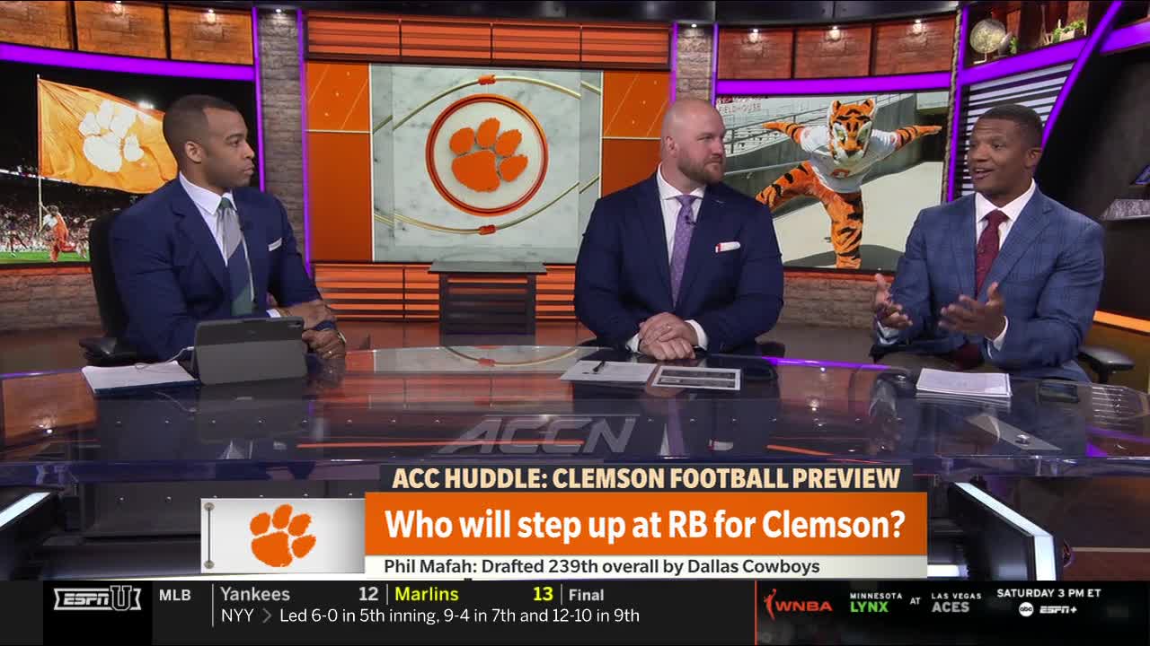 Three men in suits sit behind a desk, discussing Clemson football on ESPNU. A large screen shows the team's logo and mascot, while the on-screen text poses a question about the running back position.
