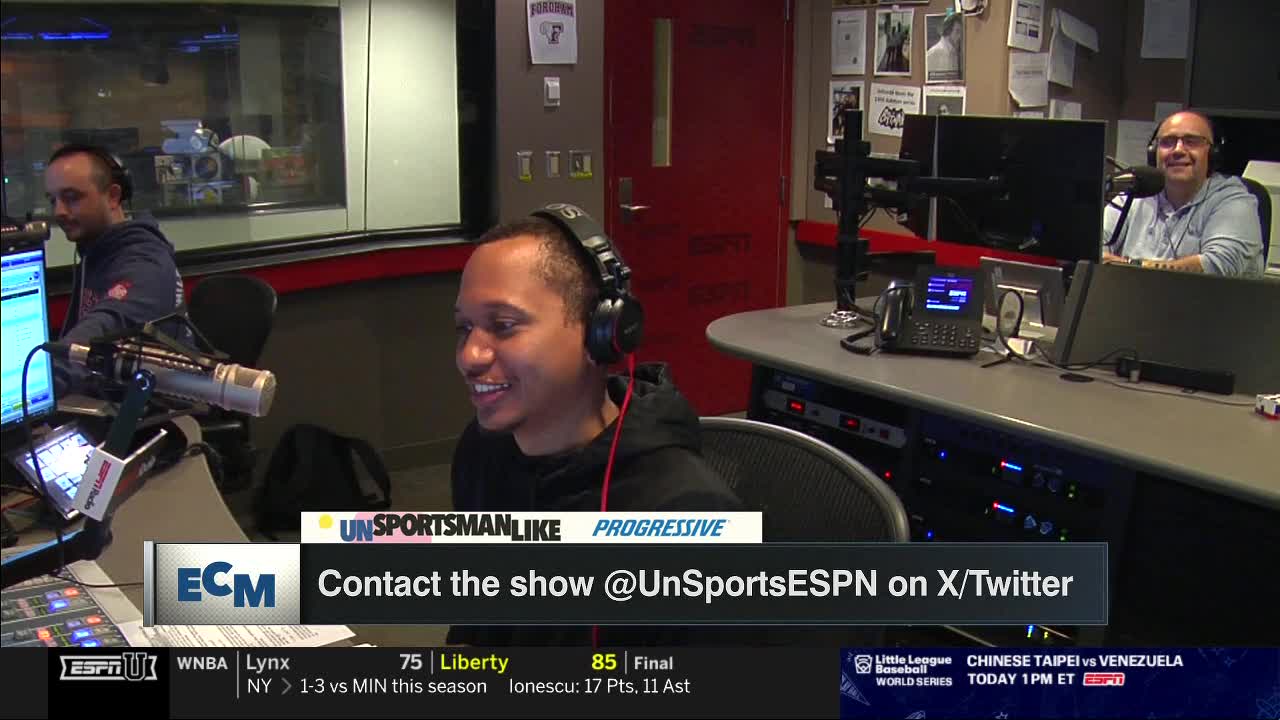 Three men are in a studio, each wearing headphones and facing microphones. The man in the center is smiling and looking to his right, while the others are also engaged in the conversation.

