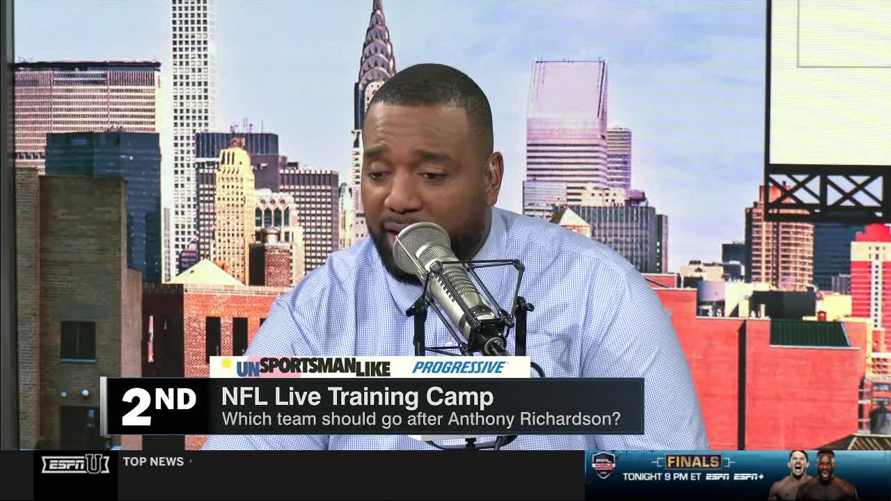 A man in a light blue shirt speaks into a microphone, his face lit by the studio lights. Behind him, a cityscape backdrop suggests the scene is part of an ESPNU broadcast about NFL training camp.
