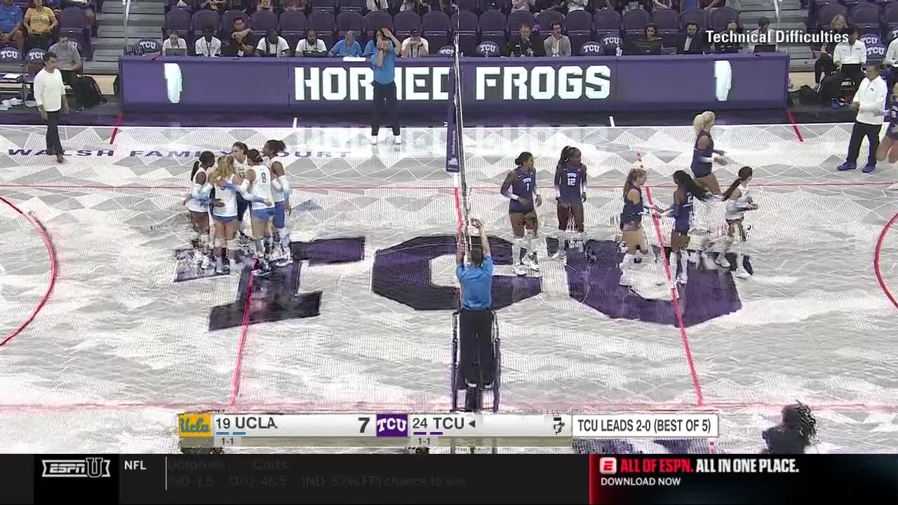 A referee adjusts the net while the TCU Horned Frogs volleyball team stands ready on their side of the court. The UCLA team huddles together on the other side, as the ESPNU broadcast shows TCU leading the match.
