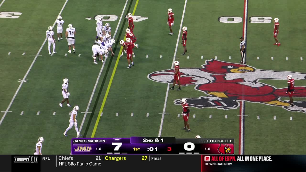 James Madison's offense is huddled near the line of scrimmage, while Louisville's defense spreads out in anticipation. The score is 7-0, and the clock shows just a second left in the first quarter on ESPNU.
