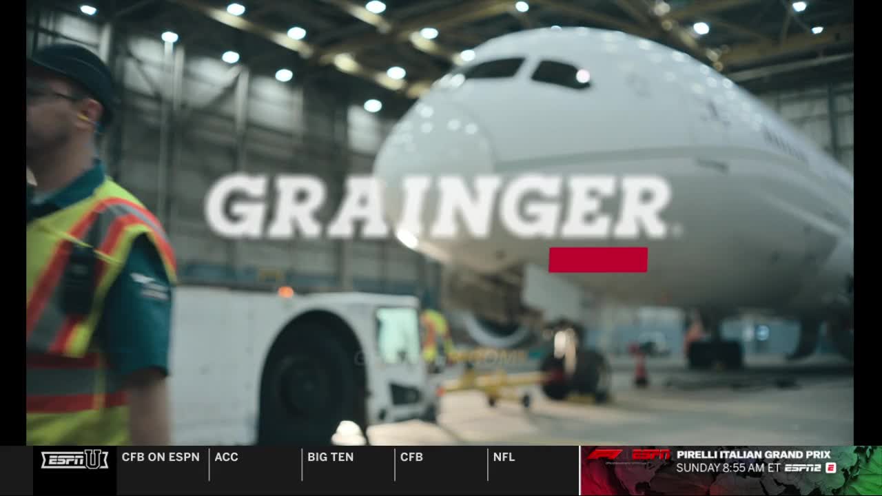 A large airplane sits inside a hangar, with "GRAINGER" superimposed over the fuselage. A worker in a safety vest walks past the nose of the plane, and the ESPN logo appears at the bottom.

