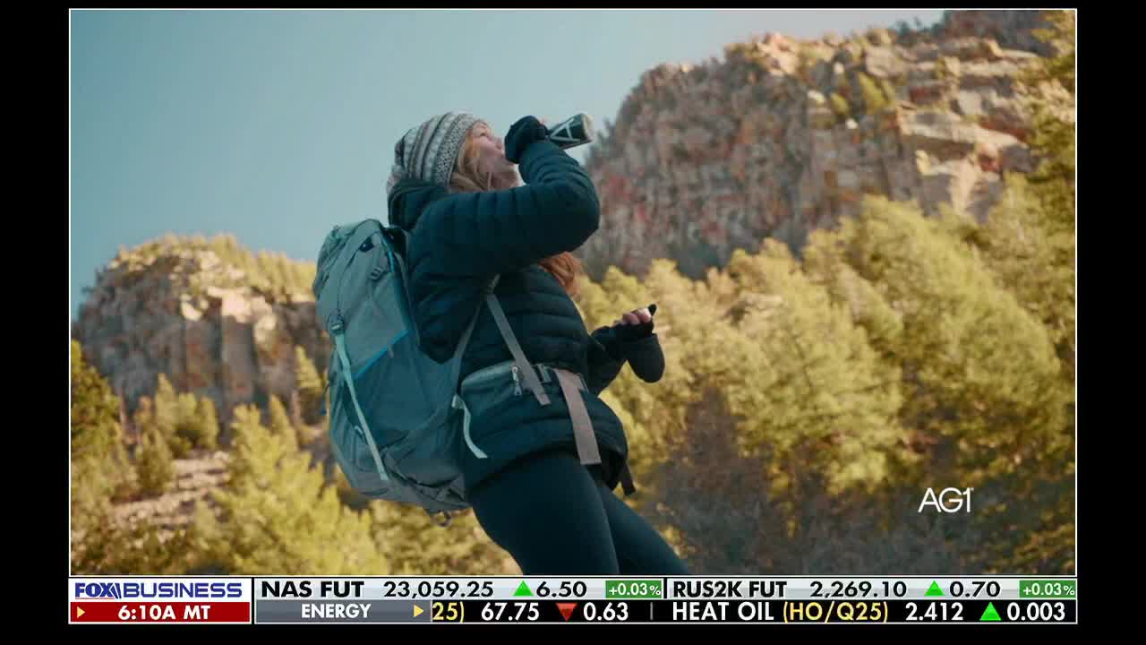 A woman, bundled up and wearing a backpack, tilts her head back to drink from a bottle. The scene is playing on Fox Business Network, with stock market data visible at the bottom of the screen.
