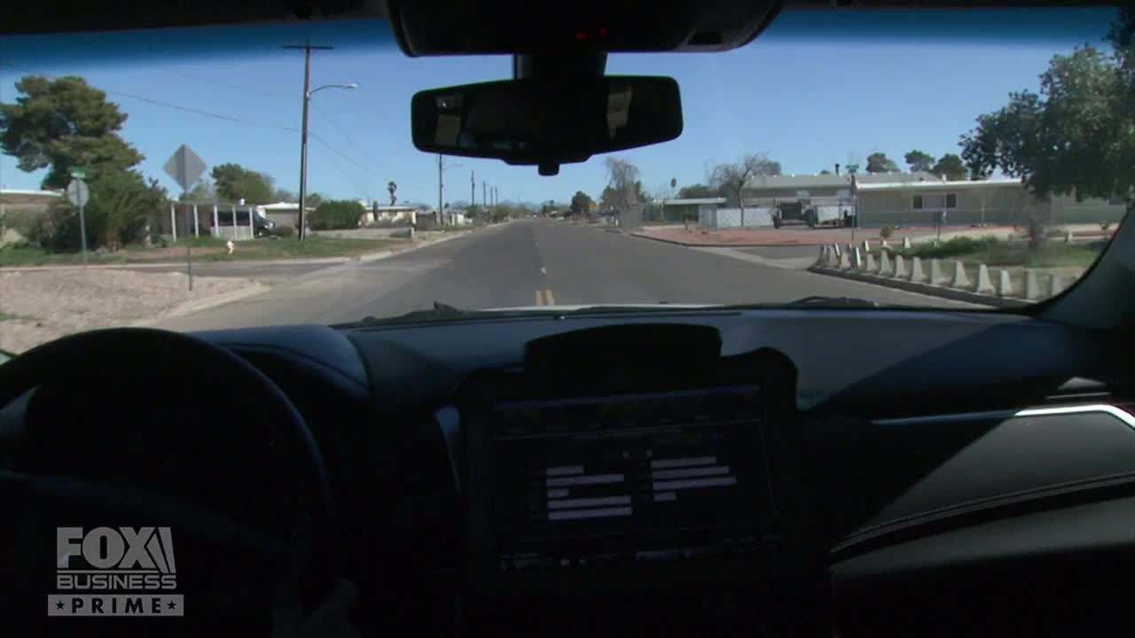 The car is driving down a residential street, with houses and trees lining the sides. The dashboard display shows data, likely related to the vehicle's performance, as seen on Fox Business Network.
