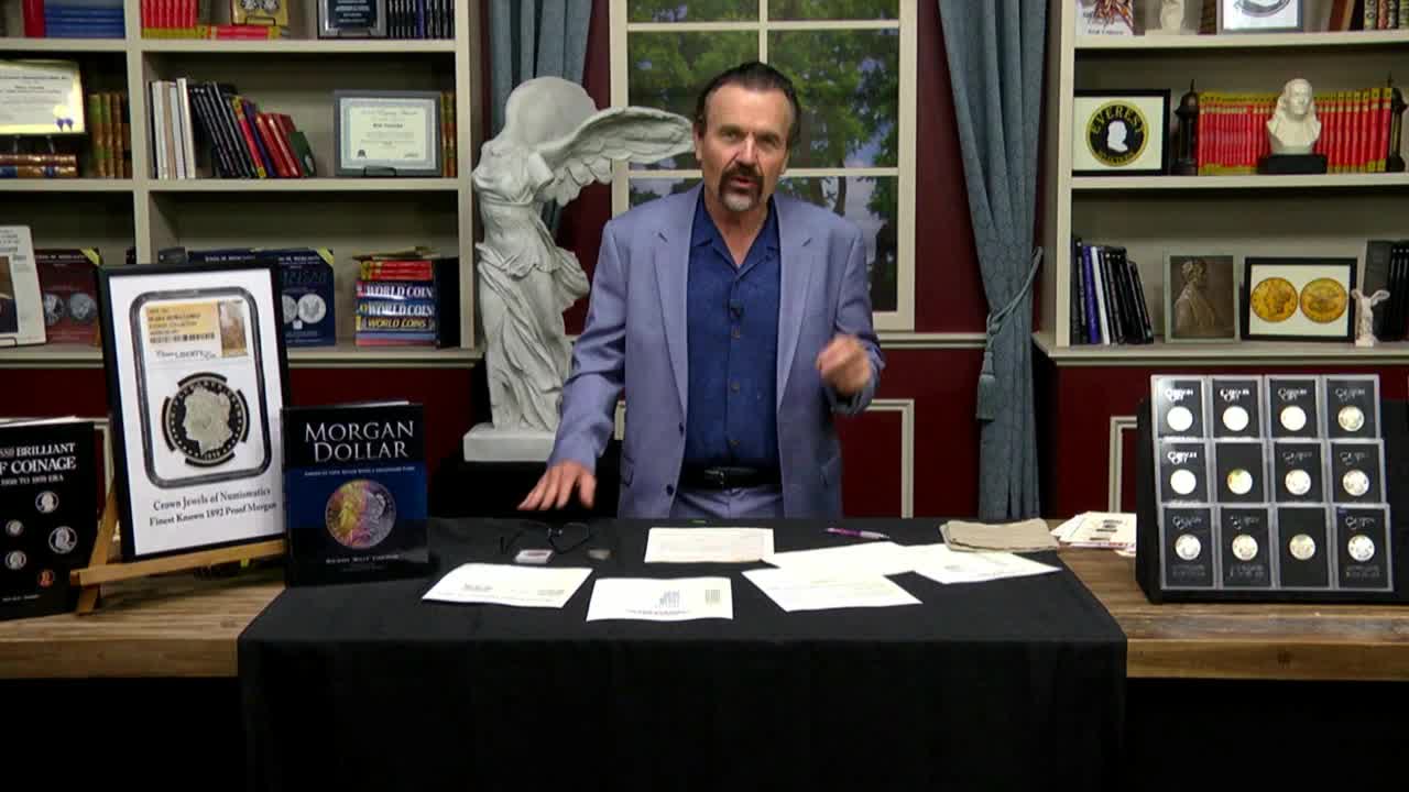 A man in a blue suit gestures toward a table covered with coins, books, and envelopes. He's standing in what looks like a television studio, possibly for Fox Business Network, with shelves of books behind him.
