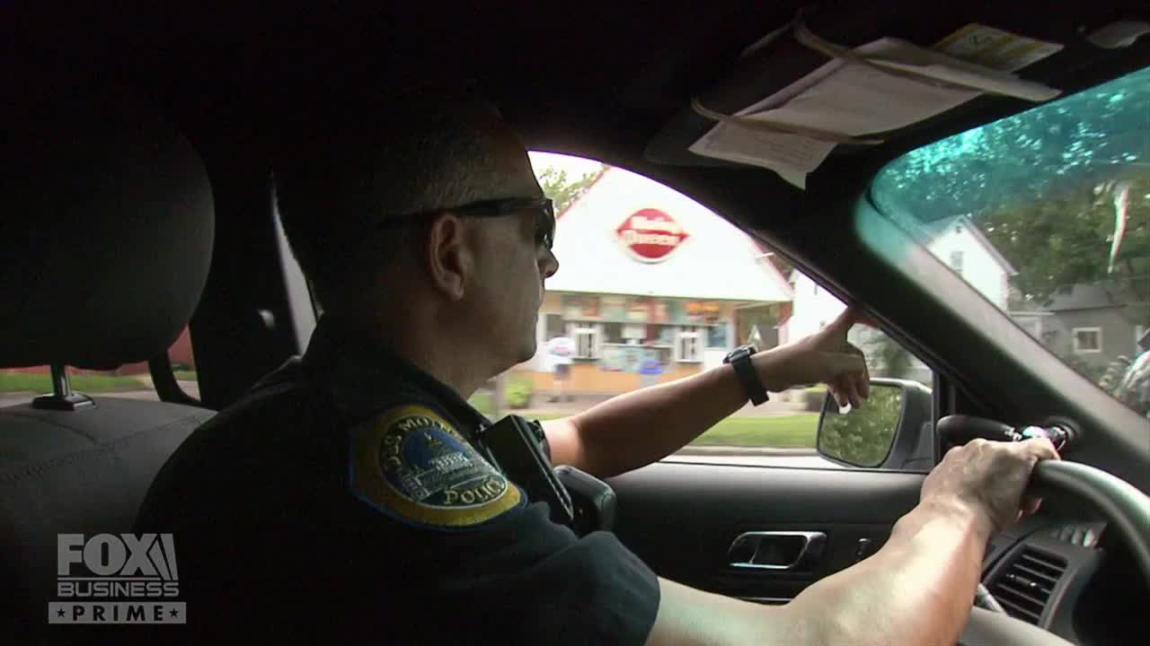 A police officer, seen from inside the car, gestures toward a Dairy Queen as they drive down a street in the United States. The Fox Business Network logo is visible in the corner.
