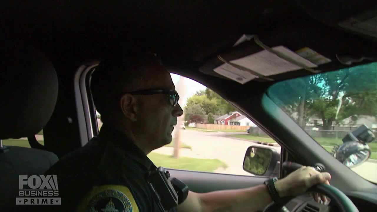 A police officer, wearing glasses, drives a patrol car down a residential street. The Fox Business Network logo is visible in the corner.
