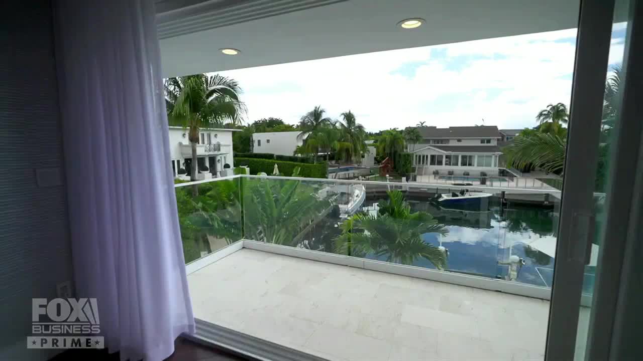 From the Fox Business Network broadcast in the United States, a white curtain gently sways near a glass balcony. Beyond, a canal reflects the sky, with boats docked near luxurious homes.
