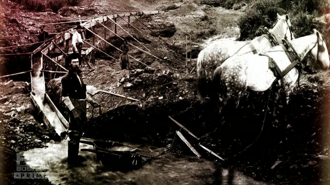 A man stands in the water, next to a sluice box, while others work on a wooden structure. Two horses, harnessed together, stand nearby.
