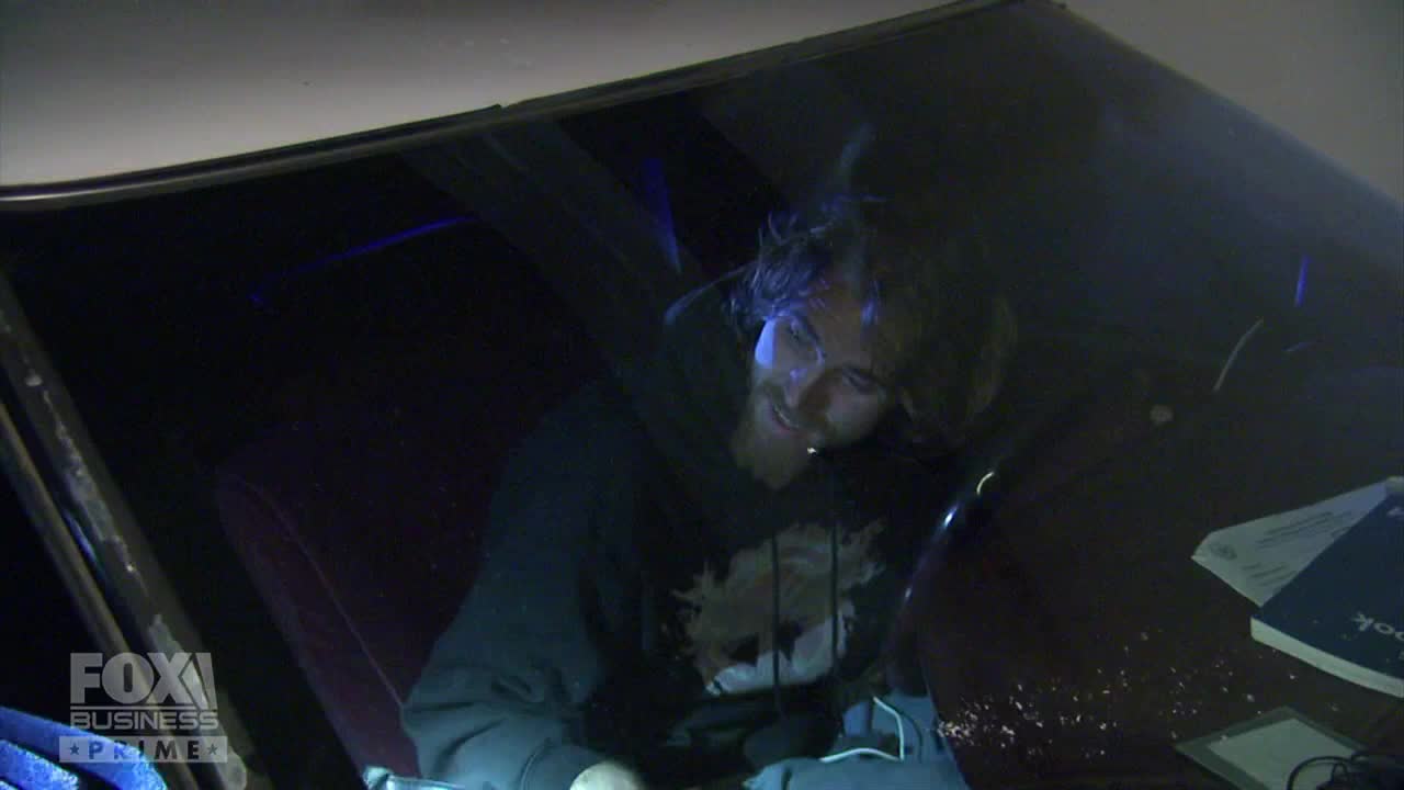 A man in a dark hoodie sits inside a car, illuminated by a blue light, as seen on Fox Business Network. He's looking down, seemingly at something on the dashboard.
