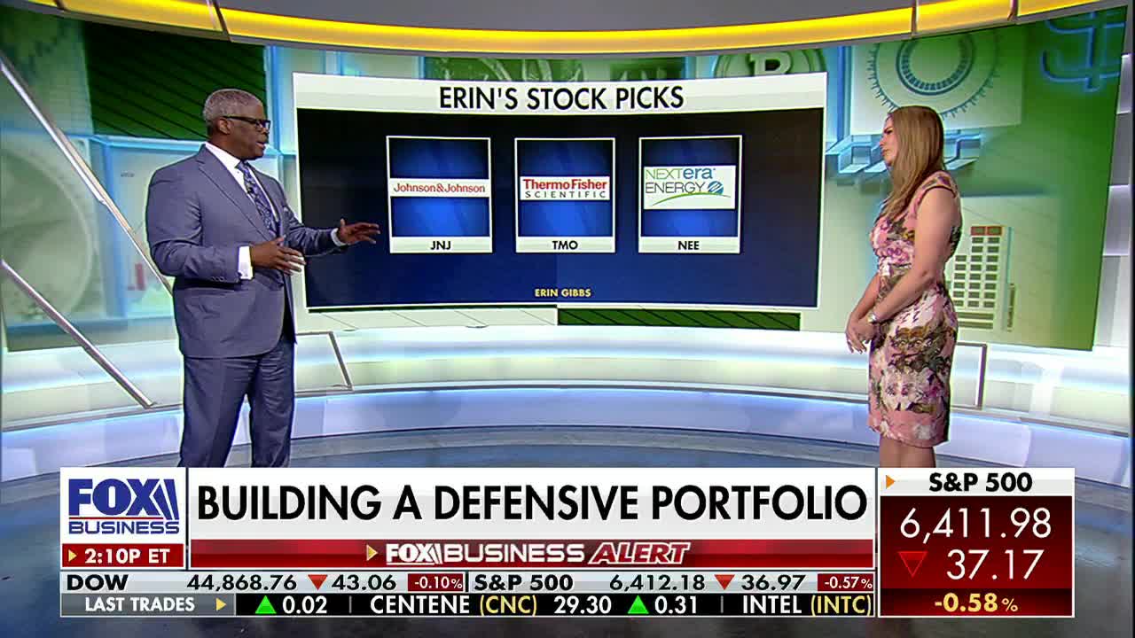On Fox Business Network, a man in a blue suit gestures toward a screen displaying stock picks. A woman in a floral dress stands nearby, listening intently.
