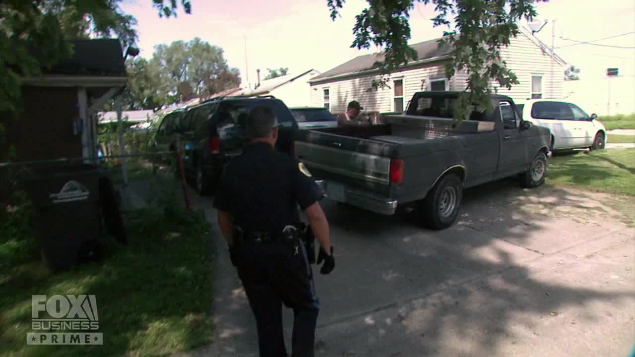 A police officer walks towards a black pickup truck parked on a driveway. Two men are visible in the bed of the truck, and a Fox Business Network logo is in the corner.
