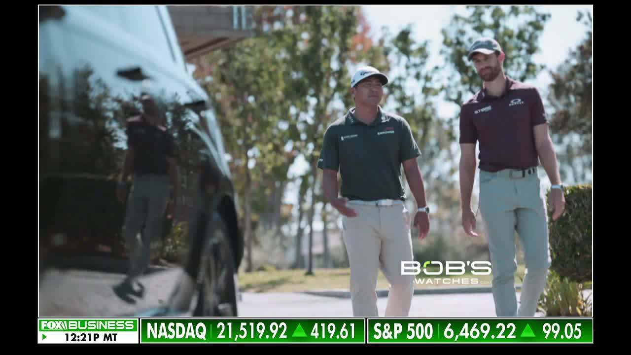 Two men in golf attire are walking toward the camera, a black vehicle parked to their left. The Fox Business Network ticker at the bottom of the screen shows the NASDAQ and S&P 500 indices.

