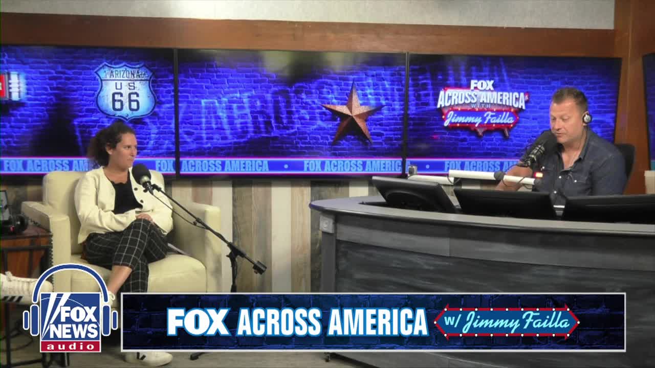 A woman in a cream-colored cardigan sits in an armchair, facing a microphone. Across from her, a man in a button-down shirt is at a desk, speaking into his own microphone.
