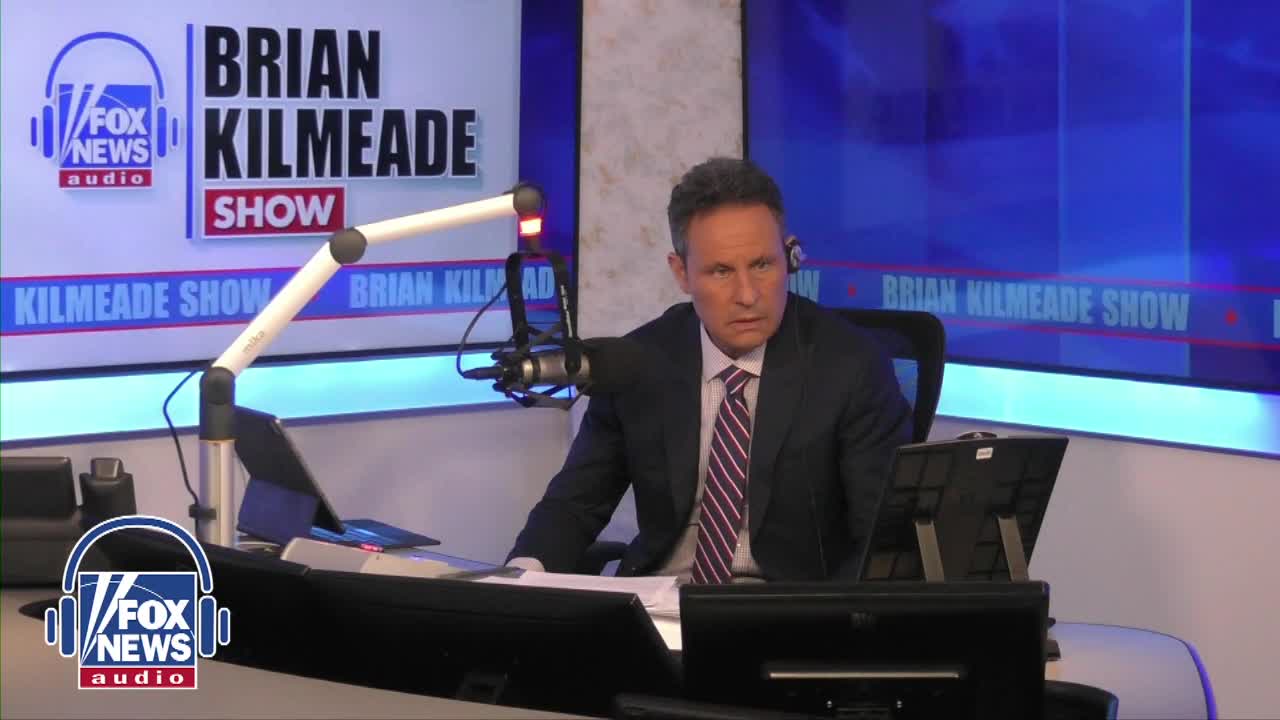 The man in the suit leans forward, his gaze fixed on something just out of frame. A microphone hangs in front of him, ready for him to speak into.