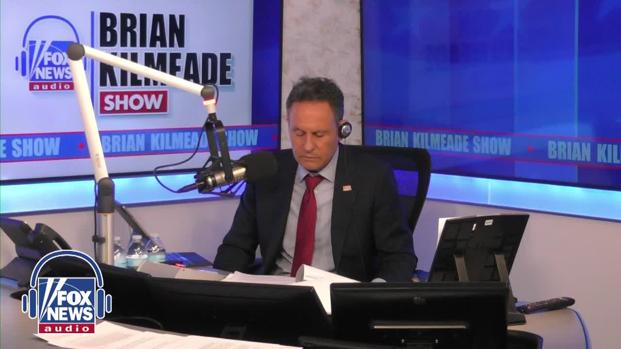 Brian Kilmeade sits at his desk, head bowed, during a broadcast of the Fox News Radio show. A microphone hangs in front of him, and the "Brian Kilmeade Show" logo is visible on a screen behind him.