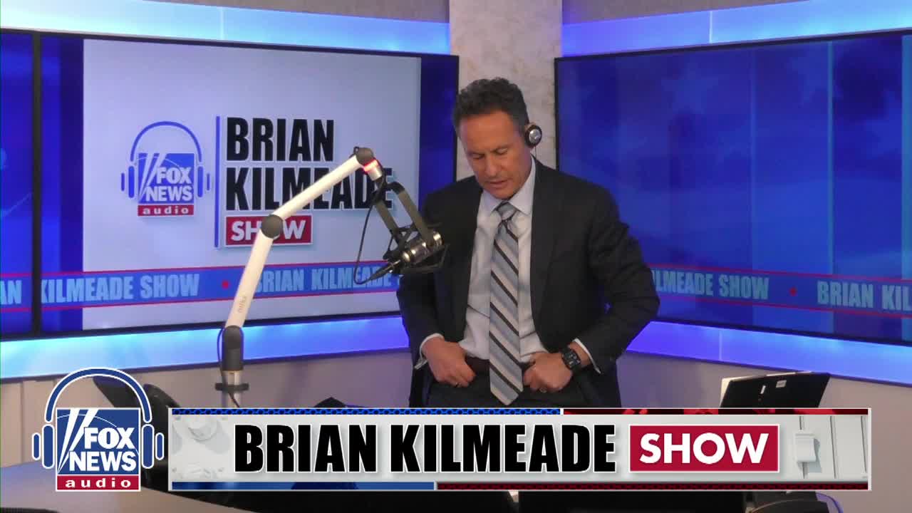 Brian Kilmeade adjusts his suit jacket in front of a microphone. The "Brian Kilmeade Show" logo is visible on a screen behind him.
