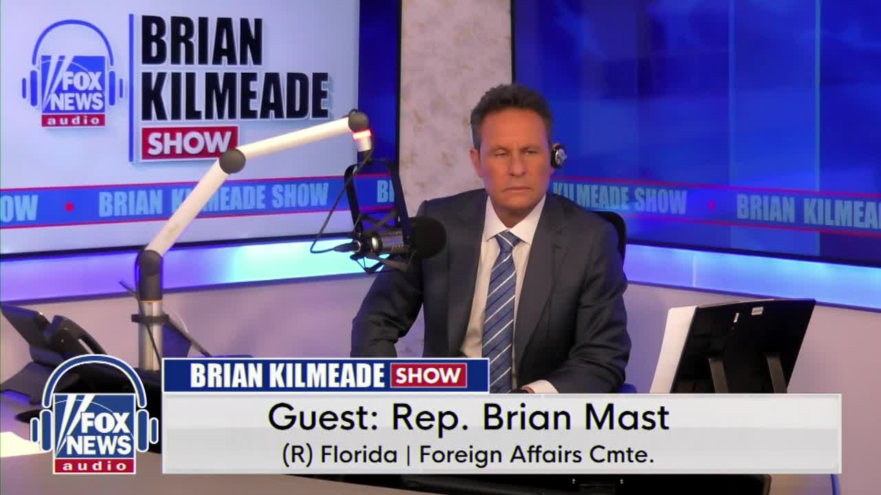 A man in a suit sits at a desk, headphones on, facing a microphone. The "Brian Kilmeade Show" logo is visible on a screen behind him.