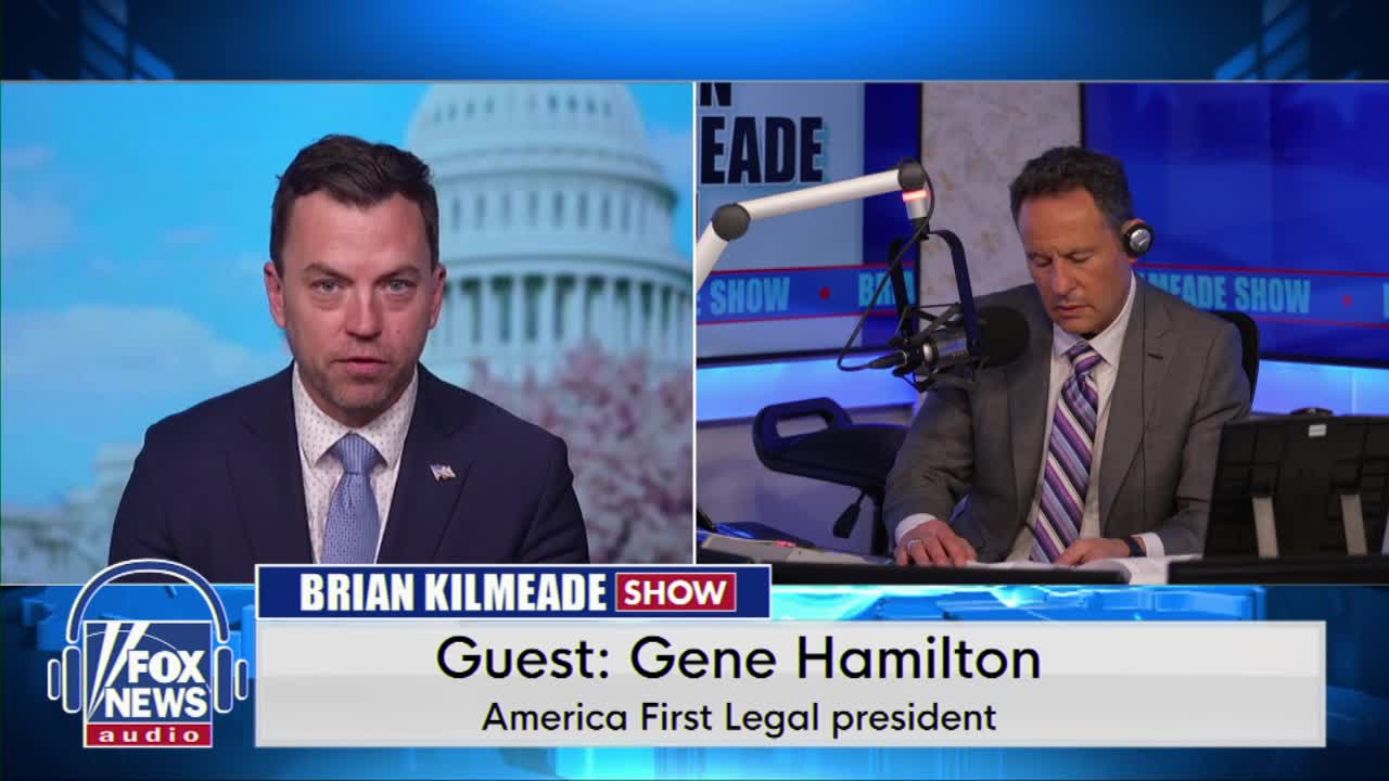 A man in a suit and tie looks directly at me, his expression serious. To his right, another man in a suit and tie with a striped tie leans over a desk, focused on papers in front of him.