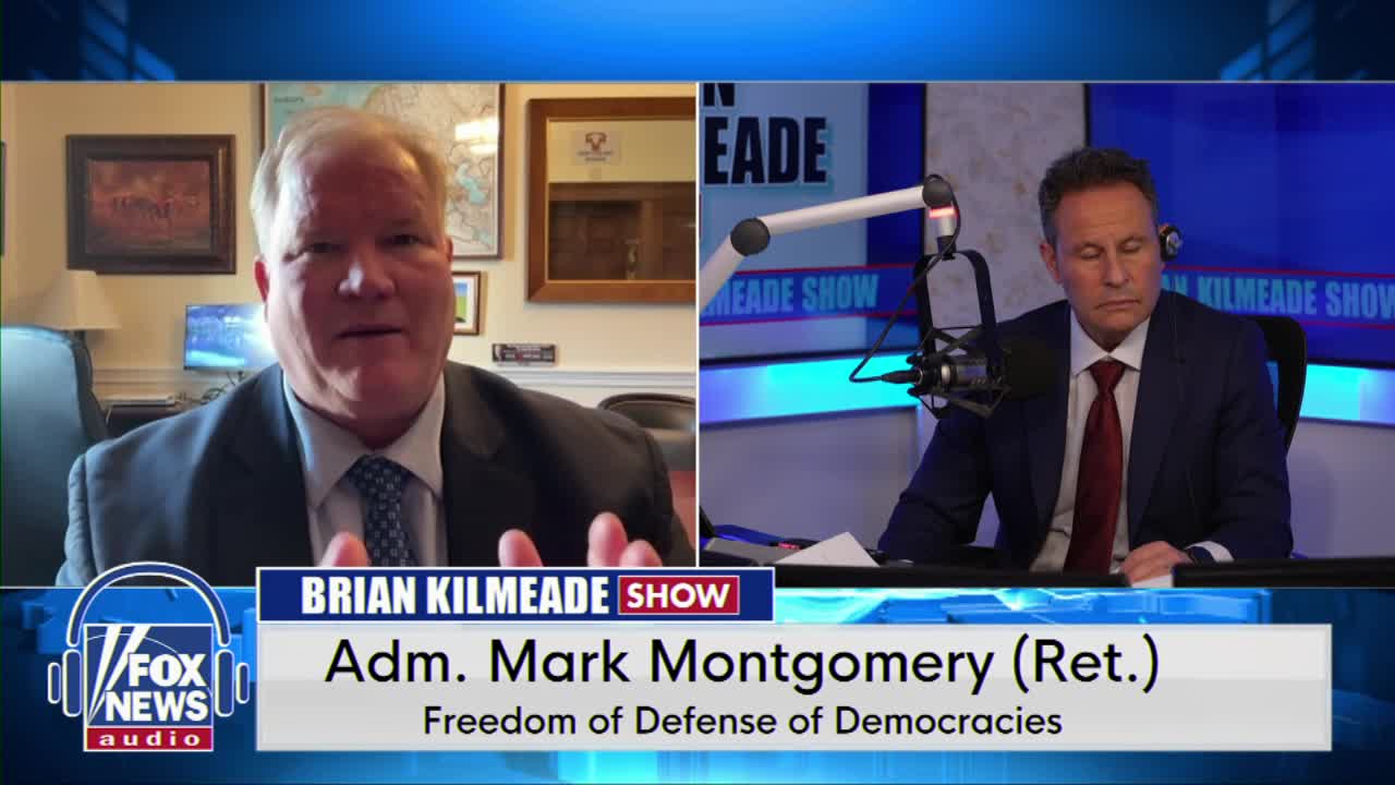 A man in a suit gestures with his hands while speaking on a video call. Across from him, another man in a suit leans forward at a microphone.