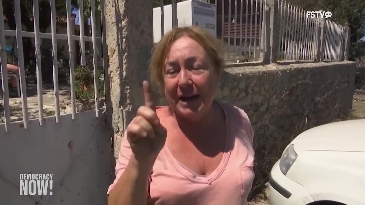 A woman, framed by a gate and a white car, gestures emphatically with her index finger. She's speaking animatedly, likely for the cameras of Free Speech TV's *Democracy Now!*
