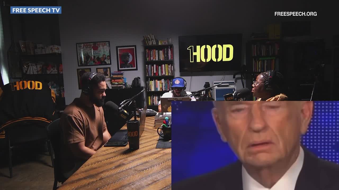 Three people sit at a table with microphones, broadcasting for Free Speech TV, while a stunned man's face is superimposed on the bottom of the screen. The word "HOOD" is displayed in large yellow letters behind them.
