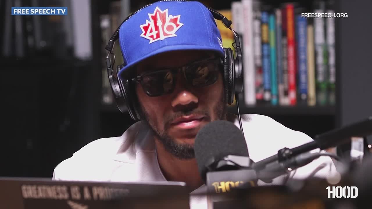 A man in a blue baseball cap and sunglasses sits at a microphone, headphones on. He's speaking on what looks like a Free Speech TV broadcast, with a bookshelf visible in the background.
